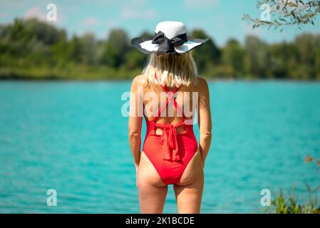 Rear view of a blonde woman with a black and white summer hat in a red swimsuit in front of a beautiful lake Stock Photo