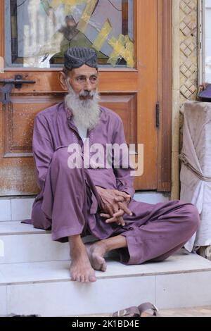 A vertical shot of a Pakistani old male with a white beard in a topi sitting barefoot on the street Stock Photo