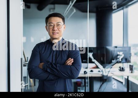 Portrait of successful Asian programmer, team leader company owner smiling and looking at camera with crossed arms, talented startup entrepreneur working inside IT office Stock Photo