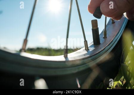 Close-up of a hand with a cap from a nipple on a bicycle wheel, in a meadow, against the background of the sky. Flat tire. Bottom view. Stock Photo