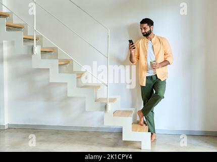 Indian man wearing earbud using smartphone standing at home. Stock Photo