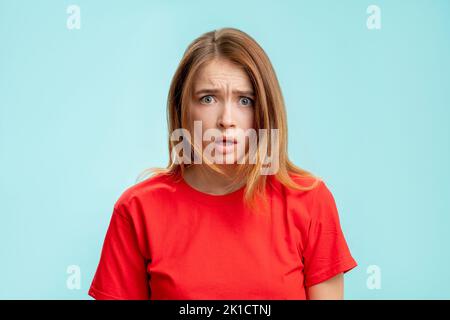 Overwhelmed woman portrait. Wtf reaction. Shocked lady in red t-shirt looking at camera questioning annoyed isolated on blue copy space background. Di Stock Photo