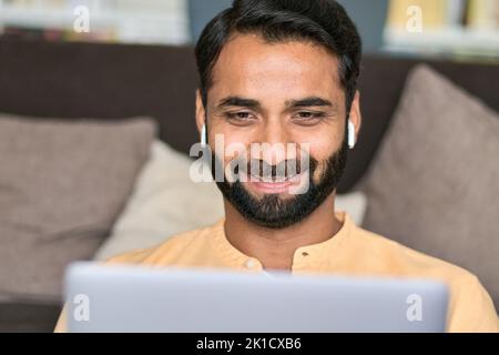 Smiling indian man sitting at home wearing earphones using laptop computer. Stock Photo
