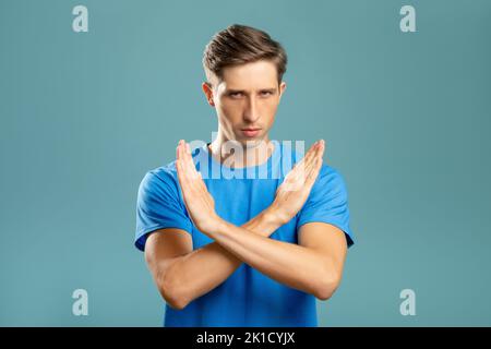 Crossed hands. Refusal gesture. Forbidden way. Stay out. Serious young man holding closed sign arms looking at camera isolated on blue. Stock Photo