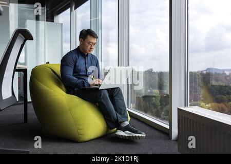 Asian young office worker sitting on soft ottoman chair, programmer using laptop for writing code and programming testing new program, man sitting near window inside office. Stock Photo
