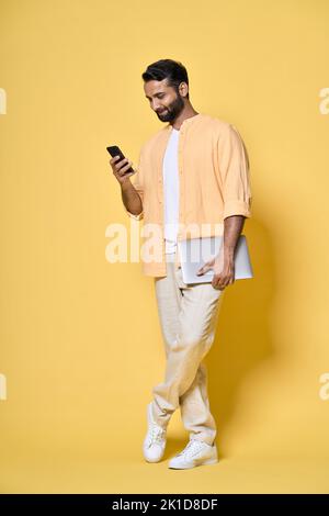 Indian man standing isolated on yellow background holding laptop using mobile. Stock Photo