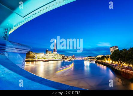 Seen from between wall opening,midway across the iconic bridge,light trail of passing river boat,the Shard and other buildings along South Bank illumi Stock Photo