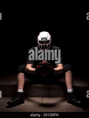 Male in American football attire sitting on a step and holding the ball with both hands. Stock Photo