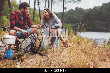 Cheerful young man and woman laughing together while camping next to the lake. Happy young couple have a great time at their campsite. Stock Photo