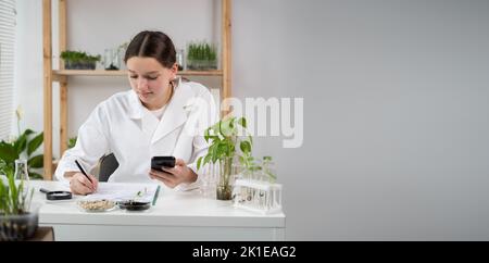 A young attractive woman scientist with protective gloves using a smartphone in the scientific bio laboratory, writing results on a clipboard. Growing Stock Photo