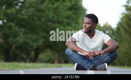 Young man in white t-shirt looks around sitting on asphalt Stock Photo