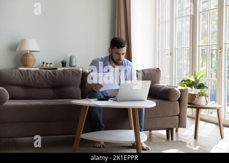 Serious focused millennial homeowner man checking paper bills Stock Photo