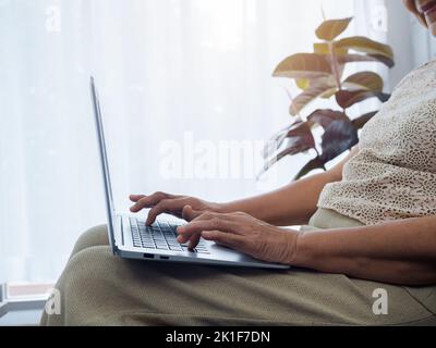 Close-up hands of senior woman working online, using laptop computer, sitting on the sofa at home. Elderly female searching something on internet. Old Stock Photo