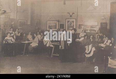 Women's tailoring or dressmaking class, early 20th century. The photograph shows young women sat in a classroom, one is working on a treadle sewing machine and the others are hand sewing. A partially completed garment is being tried on by a woman standing in the centre of the picture. Written instructions and diagrams are chalked up on blackboards and pieces of tailored garments are on display at the back Stock Photo