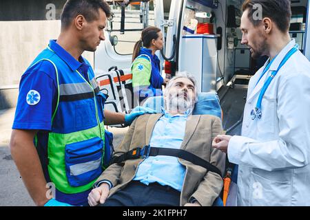Paramedic and doctor examining of senior male patient lying in ambulance gurney brought to hospital by EMT workers. Emergency medical services and car Stock Photo