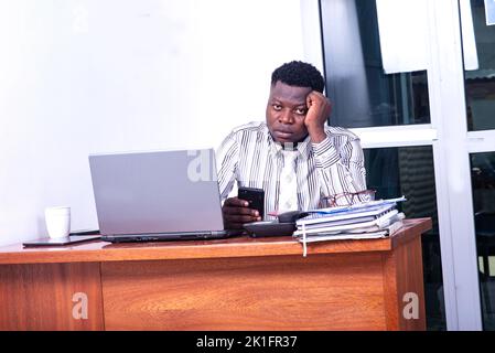 Pensive adult businessman sitting at the desk facing the computer with a mobile phone in hand and looking at the camera. Stock Photo