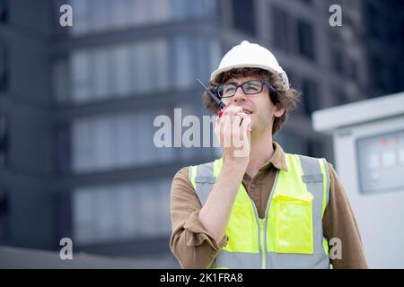 Young caucasian man using radio conversation and inspecting while building and tower all around, engineer or architect examining and maintenance for d Stock Photo