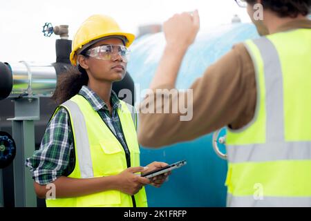 Young woman and man engineer examining pipeline and looking digital tablet in the factory, mechanic or technician inspector plumber valve and talking Stock Photo