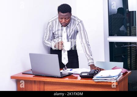 young serious businessman standing at the desk and working on the laptop while holding a cup of coffee. Stock Photo