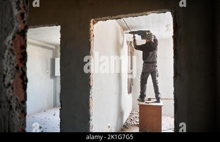 Professional strong destroyer standing on desk near gray brick wall covered with plaster and breaking it using jackhammer. Redevelopment of apartment in order to expand living space. Stock Photo