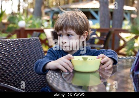 Pretty little boy sit on chair at table in cafe on summer terrace and look away. Kid of kindergarten age having dinner outdoor in forest. Children Stock Photo