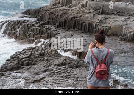 Kunashir, Russia - Juiy 31, 2022: woman tourist takes pictures of coastal rocks formed by columnar basalt in a stormy sea surf Stock Photo