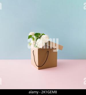 White flowers in brown gift cardboard box on blue and pink background. Flower delivery. Minimal concept. Stock Photo
