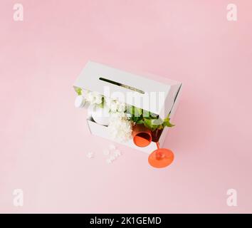 White delivery gift box with white flowers, two red drinking glasses and white drinking bottle on pink background. Stock Photo
