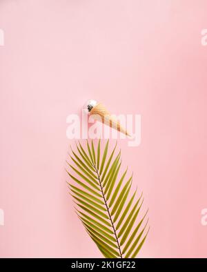 Ice cream cone with shiny disco ball sticking on the pink wall above palm leaf. Front view. Minimal concept. Stock Photo