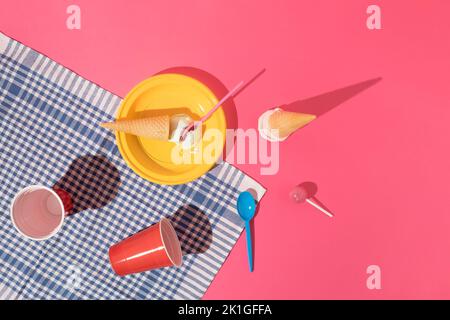 Summer picnic scene with ice cream, yellow plastic plate, red cups, blue and pink spoon, pink lollipop and blue tablecloth on pink background. Minimal Stock Photo