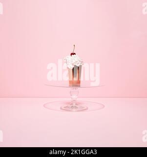 Golden paper cup filled with whipped cream and cherry on the top on glass cake stand against pink background. Summer cupcake. Minimal concept. Stock Photo