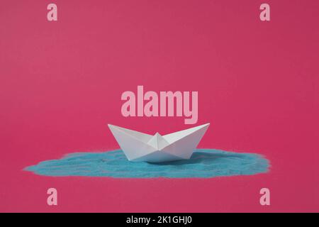 White origami paper boat floating on blue sand with pink background. Minimal concept. Stock Photo