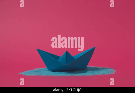 Blue origami paper boat floating on blue sand with pink background. Minimal concept. Stock Photo