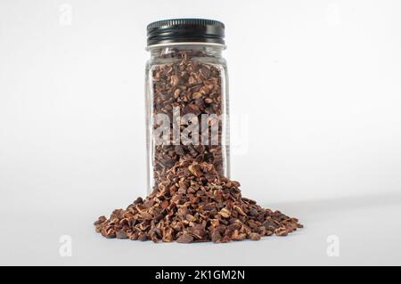 Full of organic raw cacao nibs in glass jar with black lid with a little heap in front of it. Isolated on white background. Stock Photo