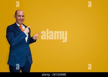 A bald Caucasian man dressed in a blue suit, white shirt and orange tie stands at one edge of the image pointing his fingers to the side where there i Stock Photo