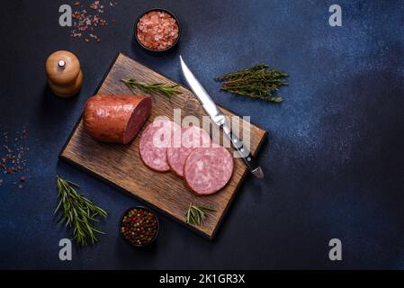 Delicious fresh smoked sausage cut with slices on a wooden cutting board against a dark concrete background Stock Photo