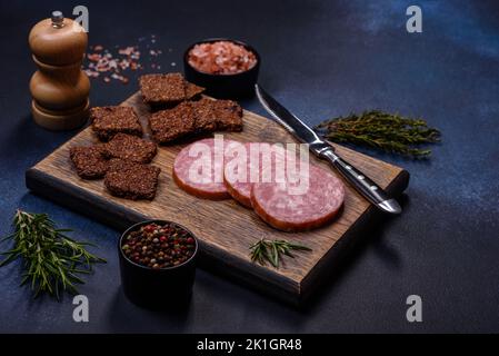 Delicious fresh smoked sausage cut with slices on a wooden cutting board against a dark concrete background Stock Photo