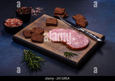 Delicious fresh smoked sausage cut with slices on a wooden cutting board against a dark concrete background Stock Photo