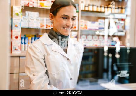 White apothecary wearing lab coat smiling while working with till at pharmacy Stock Photo