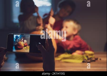 Mother using her mobile phone to take photos of her excited three kids with a halloween pumpkin they had carved. Stock Photo