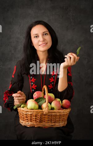 Beautiful Ukrainian woman in a black dress with a basket of ripe apples on a dark background Stock Photo