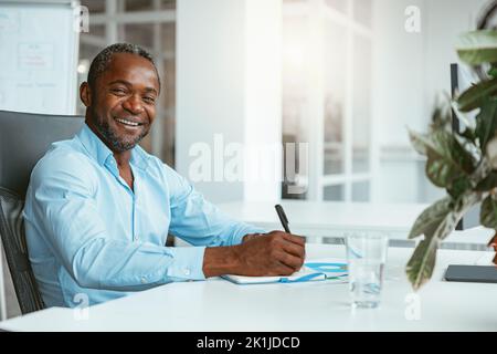 Smiling African businessman taking notes while sitting in modern office and looking at camera Stock Photo