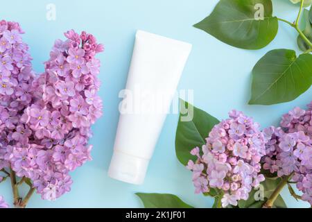 Mockup white squeeze bottle cosmetic tube, lilac flowers and leaves on blue background. Daily cream, gel, skin care, sunscreen, moisturizer. Front top Stock Photo