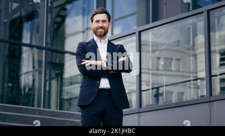 Successful confident caucasian bearded businessman in suit standing outdoors on background office building smiling proud male professional worker Stock Photo