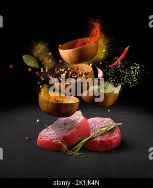 Raw beef steaks on a black stone cutting table and various spices and herbs flying with splash over black background Stock Photo