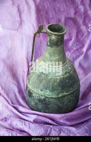Still life with an antique Moroccan bronze jug on a purple fabric. Vertical photo with vintage jug on  lilac cloth Stock Photo