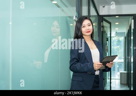 Attractive Asian businesswoman holding a digital tablet, looking at camera. Office workplace background. Stock Photo