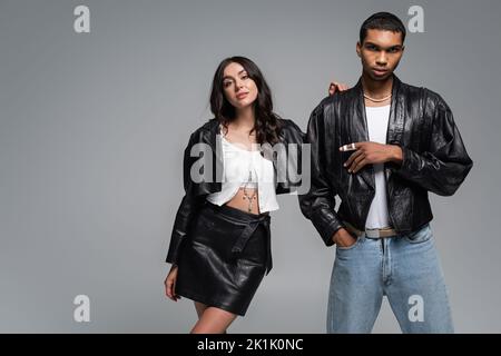 stylish african american man posing with hand in pocket near young woman in leather outfit isolated on grey,stock image
