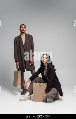 full length of stylish interracial couple in autumnal outfits posing near paper bags on grey,stock image Stock Photo