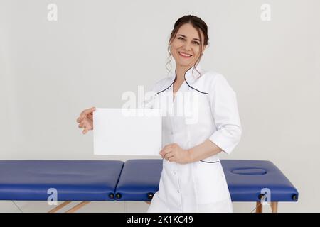 Portrait of middle-aged happy beauteous masseuse doctor woman standing near blue couch, holding blank sheet of paper. Stock Photo